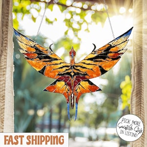 May include: A stained glass suncatcher in the shape of a stylized bird with outstretched wings. The suncatcher features orange, yellow, black, red, and blue colors. It is suspended by a chain and hangs in front of a window.