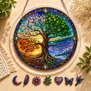 Puede incluir: Un adorno de pared redondo, estilo vidriera, que representa un árbol con cuatro estaciones. El árbol está rodeado por un borde azul con detalles de joyas. Debajo, hay broches decorativos con forma de media luna, pluma, flor, hoja, corazón, mariposa y colibrí.