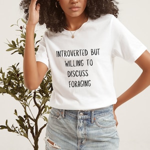 May include: White t-shirt with black text that reads "Introverted but willing to discuss foraging".