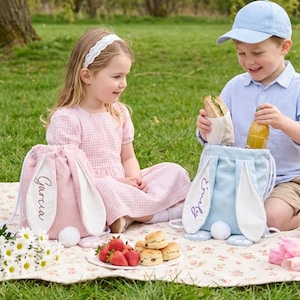 Peut inclure: Deux sacs à cordon en forme de lapin, un rose et un bleu, avec des oreilles blanches et des queues duveteuses. Le sac rose porte le nom "Garcia" brodé, et le sac bleu porte "Emily". Les sacs sont sur une couverture de pique-nique fleurie avec de la nourriture et deux enfants.