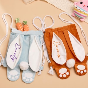 Könnte beinhalten: Zwei Hasen-Kordelzugbeutel, einer hellblau und einer rostfarben, mit weißen Hasenohren und pfotenförmigen Füßen. Auf dem hellblauen Beutel ist der Name "Emily" aufgestickt, und auf dem rostfarbenen Beutel steht der Name "Emma". Jeder Beutel hat einen weißen Kordelzug.