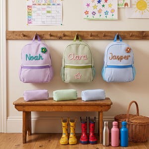 May include: Three striped backpacks in lavender, sage green, and light blue, each personalized with names Noah, Clara, and Jasper. Each bag features a matching pencil case, a small flower or star applique, and a front zipper pocket.