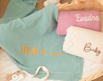 Puede incluir: Una colección de mantas para bebés en una cesta de mimbre. Las mantas son de color azul, rosa y blanco, con nombres bordados y la palabra "Baby". Una cámara de juguete y un conejito de peluche también están en la cesta.
