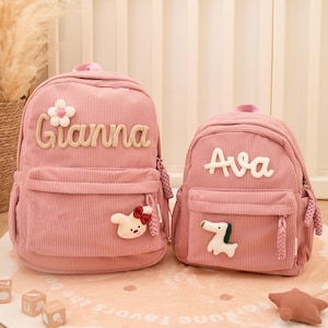 May include: Two pink corduroy backpacks, one larger and one smaller. The larger backpack has the name "Gianna" in beige script and a flower and bunny embellishment. The smaller backpack has the name "Ava" and a dog embellishment.
