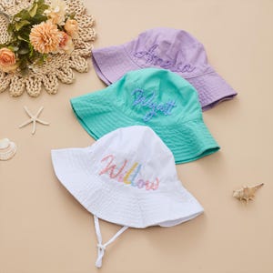 Peut inclure: Trois chapeaux de seau pour enfants, brodés de noms. Un chapeau blanc avec 'Willow' brodé en arc-en-ciel, un chapeau turquoise avec 'Wyatt' et un chapeau lilas avec 'Amelia'. Chaque chapeau a une mentonnière.