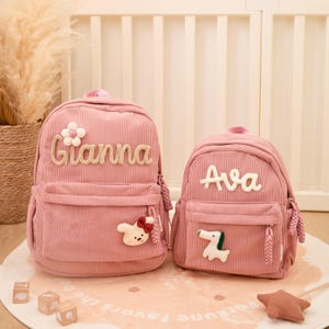Pu&ograve; includere: Due zaini rosa in velluto a coste, uno pi&ugrave; grande e uno pi&ugrave; piccolo. Lo zaino pi&ugrave; grande ha il nome "Gianna" in corsivo beige e un ornamento floreale e un coniglio. Lo zaino pi&ugrave; piccolo ha il nome "Ava" e un ornamento a forma di cane.