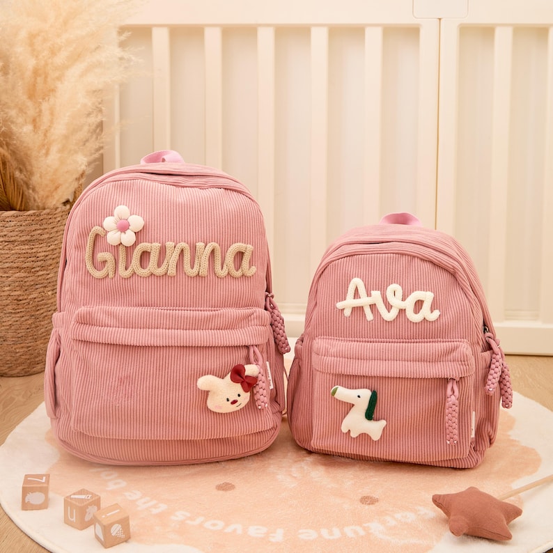 Pu&ograve; includere: Due zaini rosa in velluto a coste, uno pi&ugrave; grande e uno pi&ugrave; piccolo. Lo zaino pi&ugrave; grande ha il nome "Gianna" in corsivo beige e un ornamento floreale e un coniglio. Lo zaino pi&ugrave; piccolo ha il nome "Ava" e un ornamento a forma di cane.