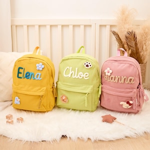 Pu&ograve; includere: Tre zaini in velluto a coste gialli, verdi e rosa, ciascuno personalizzato con un nome. Lo zaino giallo dice "Elena" in blu, quello verde "Chloe" e quello rosa "Gianna". Ognuno ha toppe decorative.