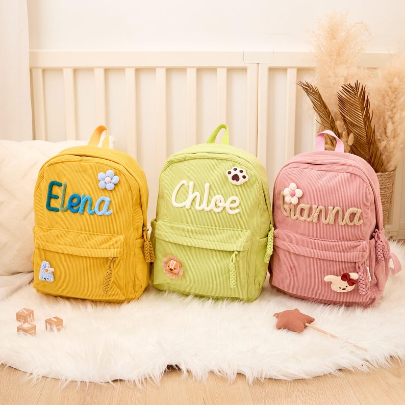 Pu&ograve; includere: Tre zaini in velluto a coste gialli, verdi e rosa, ciascuno personalizzato con un nome. Lo zaino giallo dice "Elena" in blu, quello verde "Chloe" e quello rosa "Gianna". Ognuno ha toppe decorative.