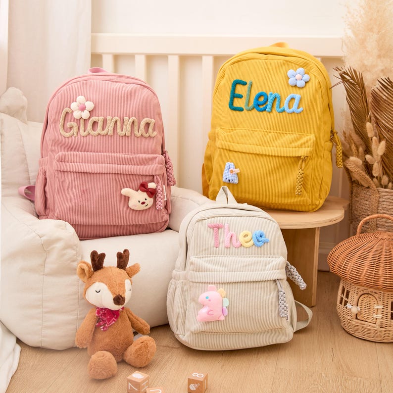 Pu&ograve; includere: Tre zaini in velluto a coste rosa, giallo e beige, ciascuno con un nome ricamato sul davanti. Lo zaino rosa ha un'applicazione a forma di fiore e coniglio. Lo zaino giallo ha un'applicazione a forma di fiore. Lo zaino beige ha un'applicazione a forma di dinosauro.