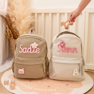 Pu&ograve; includere: Due zaini in velluto a coste, uno beige e uno grigio chiaro, ciascuno con un nome ricamato in rosa. Lo zaino beige ha un fiore rosa e una toppa a forma di orso, mentre quello grigio ha un fiore rosa e una toppa a forma di coniglio. Un giocattolo di legno viene tenuto in mano.