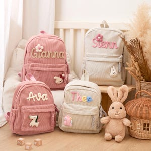 Peut inclure: Quatre sacs à dos en velours côtelé rose et beige, personnalisés avec des noms et des appliqués décoratifs. Un sac à dos rose dit "Gianna" avec une fleur, un autre dit "Ava" avec un "Z" et un troisième dit "Thee" avec un dinosaure. Un sac à dos beige dit "Sienn" avec une fleur.
