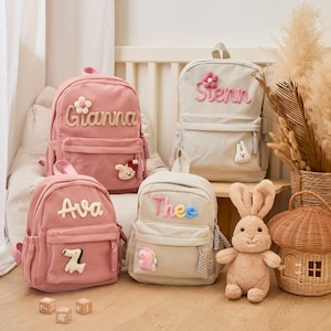 Pu&ograve; includere: Quattro zaini in velluto a coste rosa e beige, ognuno con un nome ricamato sul davanti. Gli zaini rosa hanno i nomi "Gianna" e "Ava", mentre quelli beige dicono "Stenn" e "Thee". Ogni zaino ha un piccolo ciondolo decorativo.