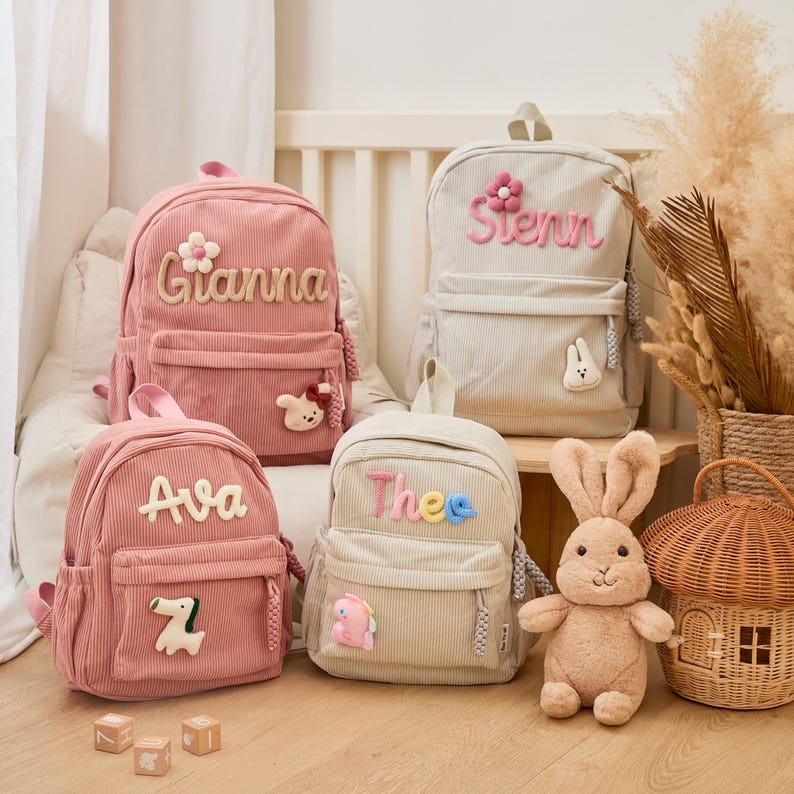 Pu&ograve; includere: Quattro zaini in velluto a coste rosa e beige, ognuno con un nome ricamato sul davanti. Gli zaini rosa hanno i nomi "Gianna" e "Ava", mentre quelli beige dicono "Stenn" e "Thee". Ogni zaino ha un piccolo ciondolo decorativo.