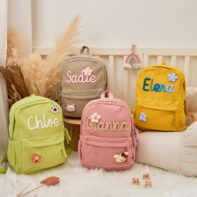 Pu&ograve; includere: Quattro zaini in velluto a coste in verde, beige, rosa e giallo. Ogni zaino ha un nome ricamato: Chloe, Sadie, Gianna ed Elena. Gli zaini hanno toppe decorative e dettagli floreali.