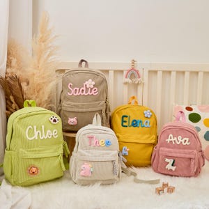 Pu&ograve; includere: Cinque zaini in velluto a coste di vari colori, ognuno personalizzato con un nome. Gli zaini sono verdi, beige, gialli, rosa e bianco sporco, con dettagli ricamati come fiori e toppe di animali. I nomi sono in un carattere giocoso.