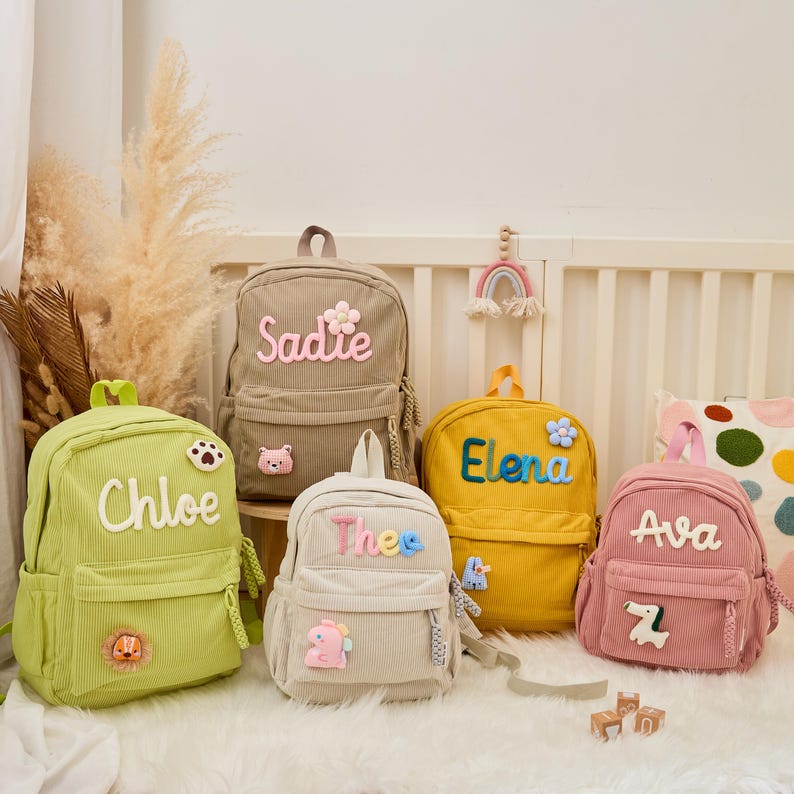 Pu&ograve; includere: Cinque zaini in velluto a coste di vari colori, ognuno personalizzato con un nome. Gli zaini sono verdi, beige, gialli, rosa e bianco sporco, con dettagli ricamati come fiori e toppe di animali. I nomi sono in un carattere giocoso.