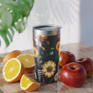 Puede incluir: Un vaso de acero inoxidable con fondo negro y un patrón floral con girasoles, rosas y otras flores en tonos amarillos, naranjas, rosas y blancos. El vaso tiene una tapa plateada.