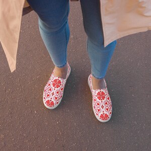 Könnte beinhalten: Ein Paar weiße Canvas-Slipper mit einem roten und rosafarbenen Herzmuster. Die Schuhe werden von einer Person getragen, die blaue Jeans und eine beige Jacke trägt.