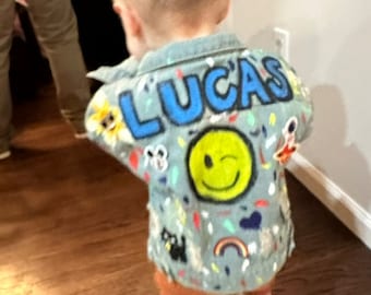 May include: A light blue denim jacket with the name "LUCAS" written in blue paint. The jacket is decorated with colorful patches, including a smiley face, a rainbow, and a flower.