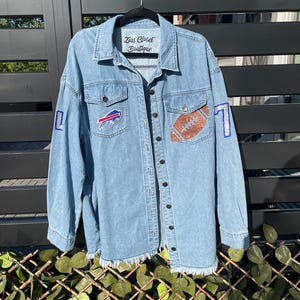 May include: A light blue denim jacket with a frayed hem. The jacket has a football, a buffalo logo, and the number "17" on it. The jacket has a label that says "Zias Closet Boutique".