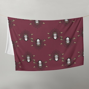 May include: A maroon fleece blanket with a repeating pattern of black and white stylized Native American headdresses with arrows. The headdresses are surrounded by a black and gold floral design.