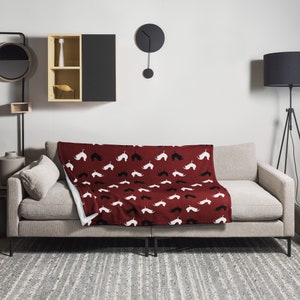 May include: A burgundy throw blanket with a repeating pattern of white and black mustache silhouettes. The blanket is draped over a light gray sofa in a living room setting. The blanket's design adds a touch of whimsy to the room's decor.