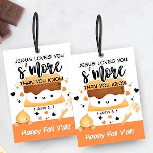 Può includere: Due etichette bianche con anelli neri presentano una grafica di s'more e il testo "JESUS LOVES YOU S'MORE THAN YOU KNOW" con "Happy Fall Y'all!" in arancione. Le etichette sono circondate da ingredienti per s'mores.