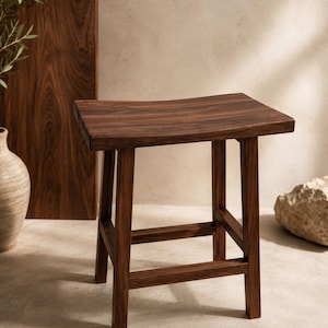 Puede incluir: Un taburete de madera marrón oscuro con un asiento rectangular curvado y cuatro patas rectas conectadas por barras de soporte horizontales. La veta de la madera es visible en toda la superficie, mostrando un tono profundo y rico.