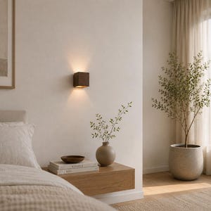 Könnte beinhalten: Ein modernes Schlafzimmer mit einer quadratischen, braunen Wandleuchte, die warmes Licht ausstrahlt. Ein Nachttisch aus Holz trägt eine Vase mit Grün, Bücher und eine Schale. Eine große Topfpflanze und ein Fenster mit Vorhängen vervollständigen die Szene.
