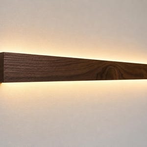 May include: A rectangular, dark brown wooden wall sconce with a warm, diffused light emanating from the bottom edge. The wood grain is visible, and the light creates a soft glow against the wall. This modern fixture is designed for interior lighting.