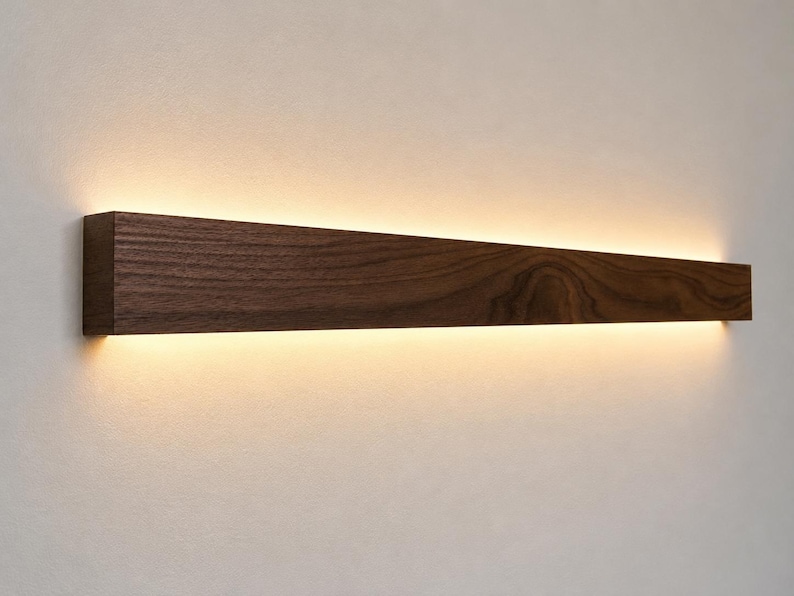 May include: A rectangular, dark brown wooden wall sconce with a warm, diffused light emanating from the bottom edge. The wood grain is visible, and the light creates a soft glow against the wall. This modern fixture is designed for interior lighting.