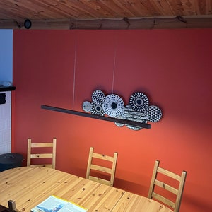 May include: A dining room scene featuring a wooden table and chairs. Above the table, a decorative light fixture with black and white patterned circular elements hangs against a red wall. The ceiling is made of wooden planks.