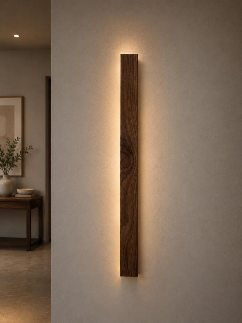May include: A vertical, rectangular wooden wall sconce with a warm, glowing light emanating from the center. The sconce is made of dark brown wood and is mounted on a neutral-colored wall. The design is minimalist and modern.
