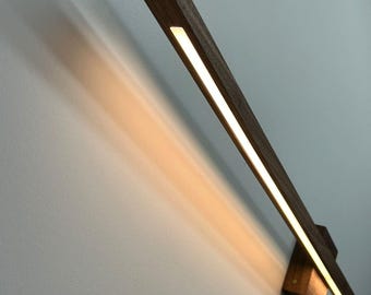 Puede incluir: Aplique de pared moderno de madera oscura con una fuente de luz LED lineal. La lámpara está montada en una pared y emite una luz cálida. El diseño es minimalista, adecuado para interiores modernos.
