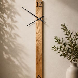 May include: A vertical wooden wall clock with black hands and the numbers 12 and 6. The clock is made of light-colored wood and has a minimalist design. The clock is mounted on a neutral-colored wall. A vase with greenery is in the background.