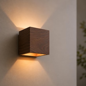 Peut inclure: Applique murale carrée en bois brun foncé, dégageant une lumière chaleureuse. Fixée sur un mur neutre, elle projette la lumière vers le haut et vers le bas. Le design est moderne et minimaliste, adapté à divers styles d'intérieur.
