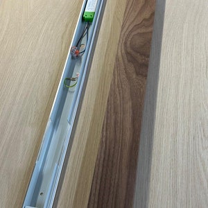 May include: An overhead lighting fixture with a silver metal housing and a dark brown wooden panel. The fixture contains electrical components, including a green-colored power supply. The wooden panel is rectangular.
