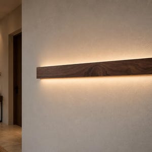 May include: A rectangular, dark brown wooden wall light fixture with a warm, yellow glow emanating from the bottom. The wood grain is visible. The light is mounted on a light beige wall.