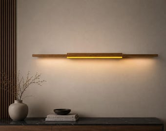May include: A modern, rectangular wooden wall sconce with a warm, yellow light. The sconce is made of light brown wood and is mounted horizontally on a neutral-colored wall. The design is minimalist and elegant, suitable for interior decor.