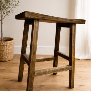 Könnte beinhalten: Ein rustikaler Holzstuhl mit rechteckiger Sitzfläche und vier stabilen Beinen. Der Hocker besteht aus hellbraunem Holz mit sichtbarer Maserung und natürlicher Oberfläche. Das Design ist schlicht und funktional, passend für verschiedene Einrichtungsstile. Höhe: ca. 45 cm.