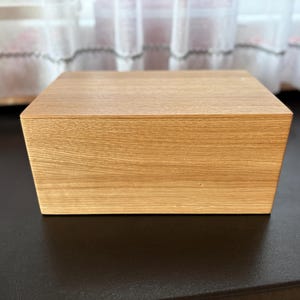 Könnte beinhalten: Eine hellbraune Holzkiste mit quadratischer Form. Die Kiste hat eine natürliche Holzmaserung und steht auf einer dunklen Oberfläche. Die Kiste ist ein schlichtes, minimalistisches Design.