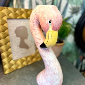 Könnte beinhalten: Eine rosa Flamingo-Figur aus Keramik mit gelbem Schnabel und schwarzen Akzenten. Der Flamingo hat einen langen, gebogenen Hals und eine kleine Öffnung oben. Die Figur ist ein dekorativer Artikel.