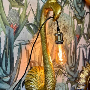 Puede incluir: Una lámpara de pared dorada con forma de flamenco, con un cable negro y una bombilla de estilo vintage. La lámpara está montada en una pared con papel pintado de estampado botánico.