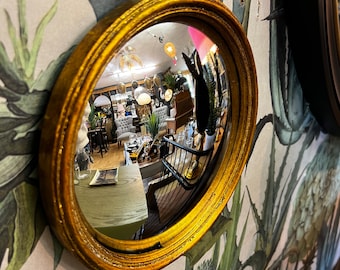 May include: A gold-framed round mirror with a convex lens reflecting a room with furniture and plants. The mirror is hanging on a wall with a floral pattern.