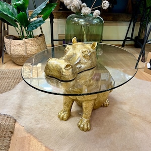 May include: A gold-coloured hippo-shaped coffee table with a clear glass top. The hippo is in a sitting position. The table is placed on a light-coloured rug. The table's design is a unique home decor item.