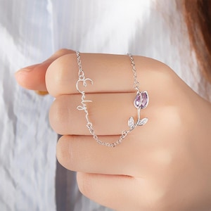 May include: A silver necklace featuring the name "Evelyn" in cursive script. The necklace includes a tulip pendant with a purple gemstone center and small, sparkling accents. The chain is delicate and the overall design is elegant.