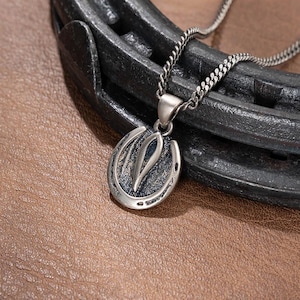 Pode incluir: Colar com pingente de ferradura prateado. O pingente apresenta um design de ferradura com uma forma central estilizada de folha ou chama. O colar é exibido em uma superfície marrom semelhante ao couro, com uma ferradura de metal escuro ao fundo.