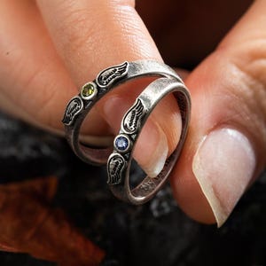 Op de afbeelding: Twee zilveren ringen met vleugelontwerpen en edelstenen. E&eacute;n ring heeft een groene edelsteen, de andere een blauwe edelsteen. De ringen worden aan een vinger gedragen en tonen de ingewikkelde details van de sieraden.