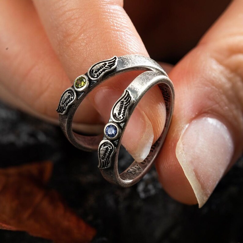 Op de afbeelding: Twee zilveren ringen met vleugelontwerpen en edelstenen. E&eacute;n ring heeft een groene edelsteen, de andere een blauwe edelsteen. De ringen worden aan een vinger gedragen en tonen de ingewikkelde details van de sieraden.